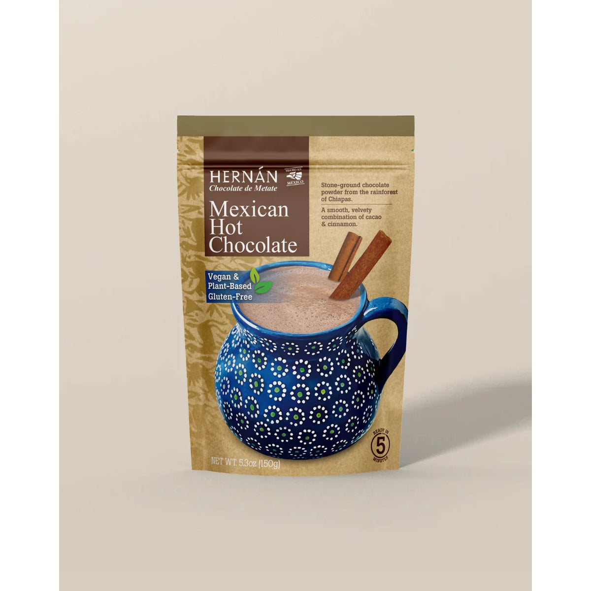A stand-up pouch of Hernán Chocolate de Menos Mexican Hot Chocolate, featuring a kraft paper design with a blue mug illustration and cinnamon sticks, set against a solid beige background.