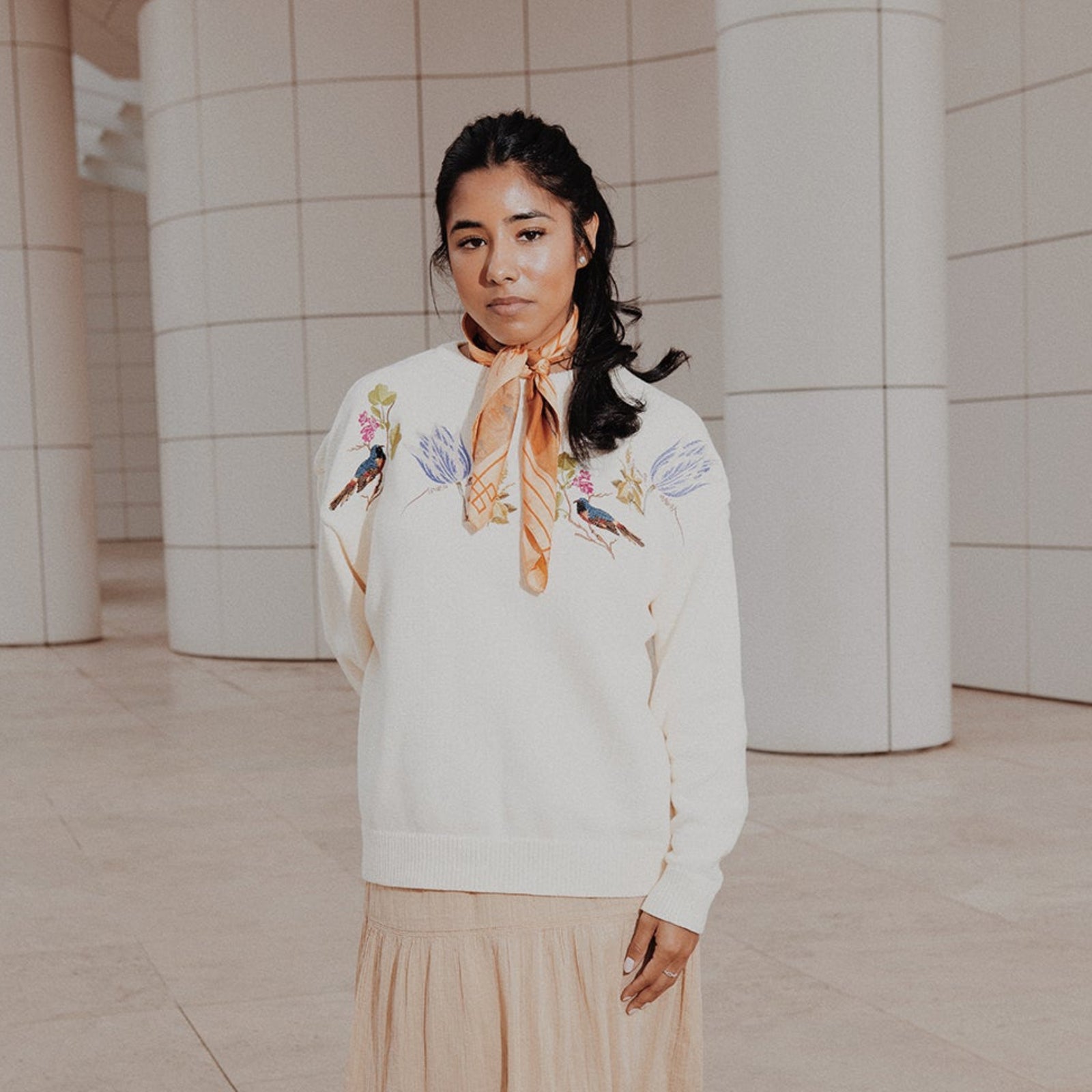 Woman in a white sweater with floral patterns and beige skirt standing in a modern architectural setting.