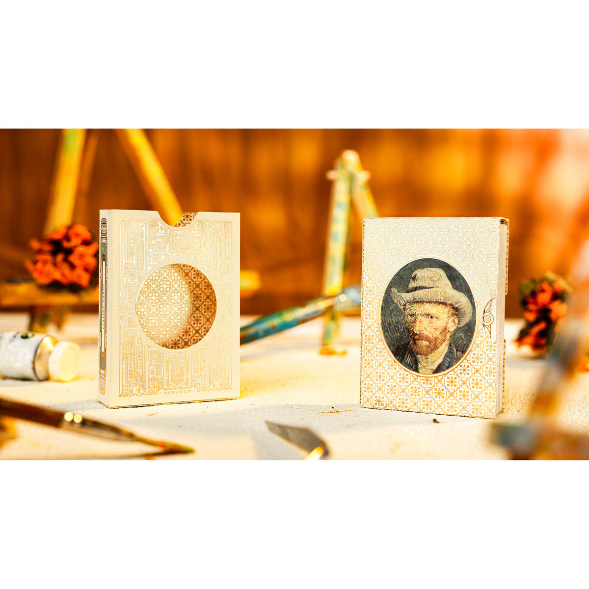 Two decorative card sleeves or small boxes standing upright on a tabletop, one with a circular cutout and one featuring a portrait of Vincent van Gogh, set against a warm, softly blurred background with paintbrushes and art supplies.