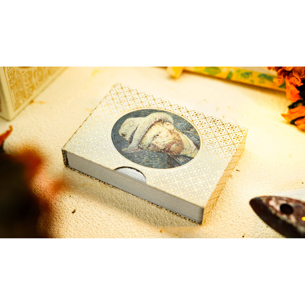 A small decorative box or package with a circular printed illustration on the lid, photographed on a light textured surface with warm lighting. The product is slightly angled, and part of another object is visible at the left edge, with a dark blurred shape at the bottom right.