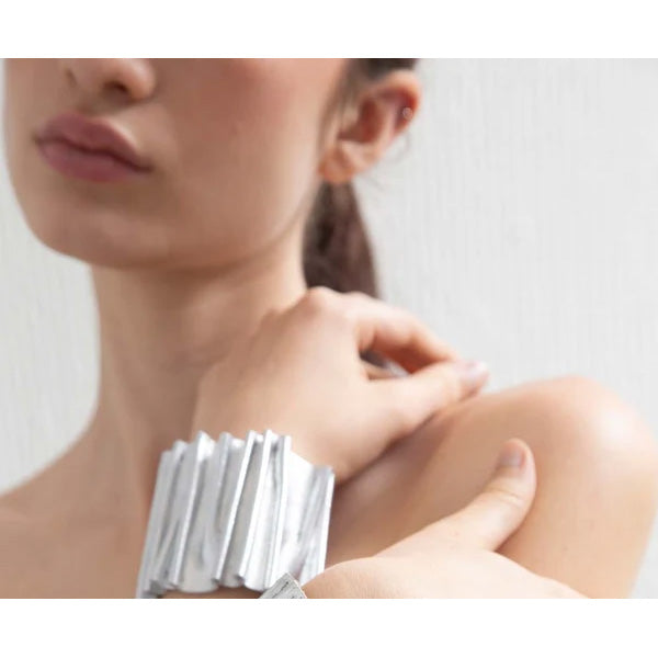 A close-up lifestyle shot of a model wearing a wide, chunky silver metallic cuff bracelet on her wrist, with her hand resting on her shoulder against a light gray background.