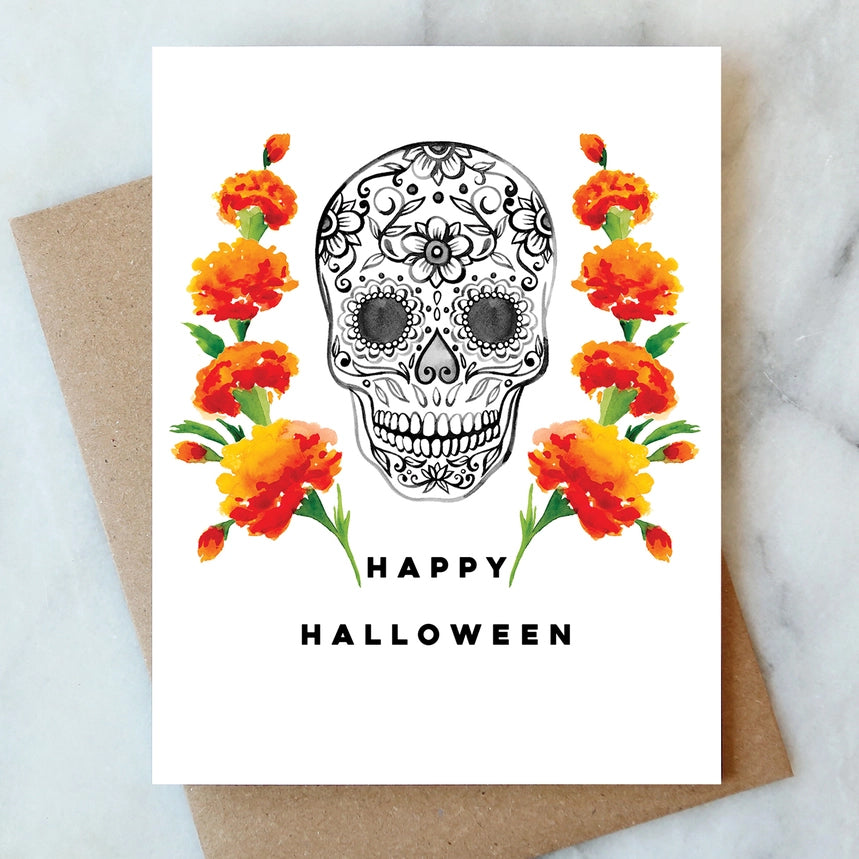 A white greeting card with a black-and-white sugar skull and orange marigold flowers, placed on top of a brown kraft envelope on a light marble surface. The card reads “HAPPY HALLOWEEN.