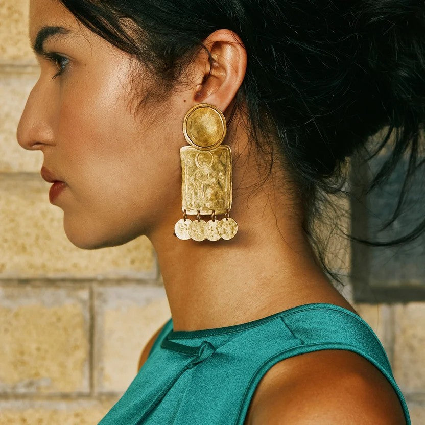 A pair of chunky, handcrafted silver-tone earrings with embossed figures and circular hammered dangles, displayed on a white background.