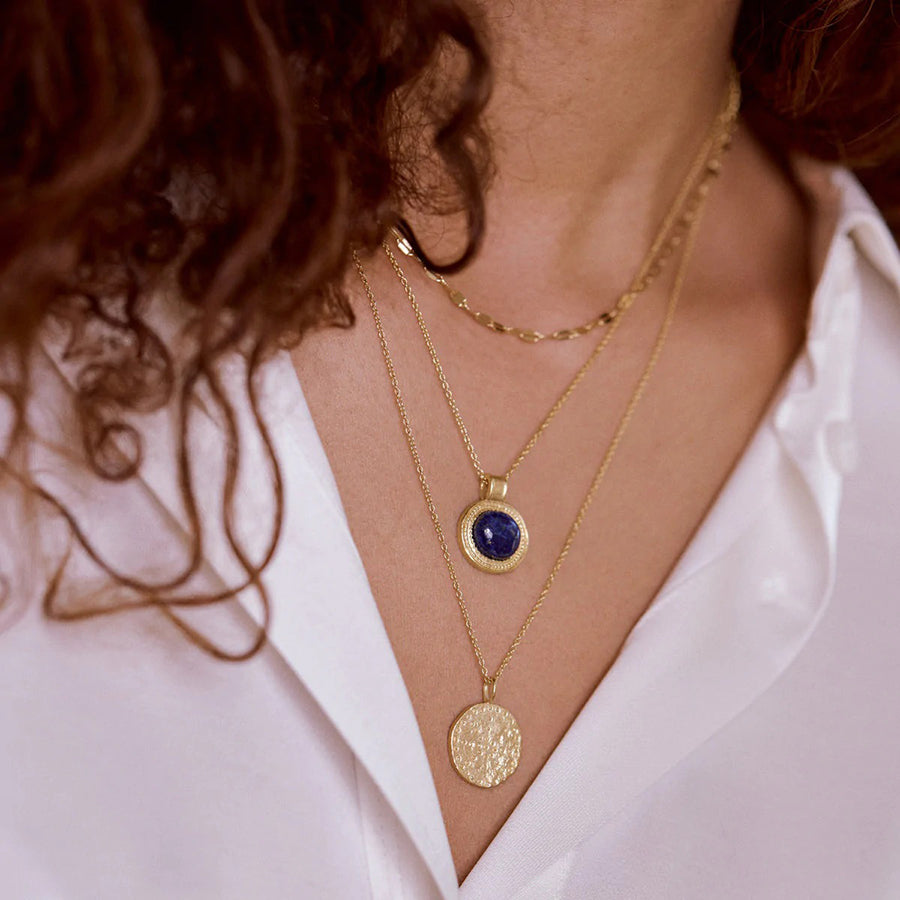 A gold-plated necklace with a round blue lapis lazuli stone set in a gold frame, displayed on a white background.