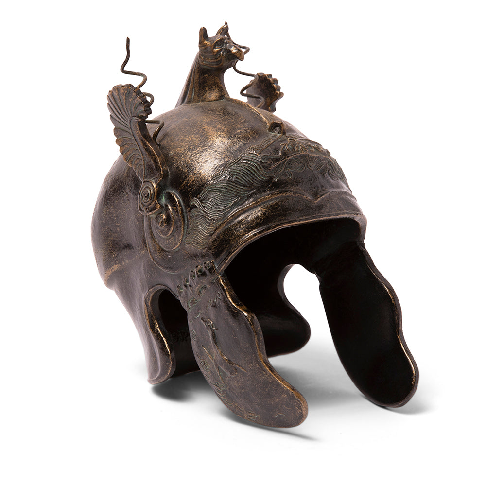 A bronze-colored reproduction of an ancient Greek Chalcidian helmet with elaborate decoration, featuring a griffin's head on the peak, wings, and engraved details on the brow and cheekpieces.