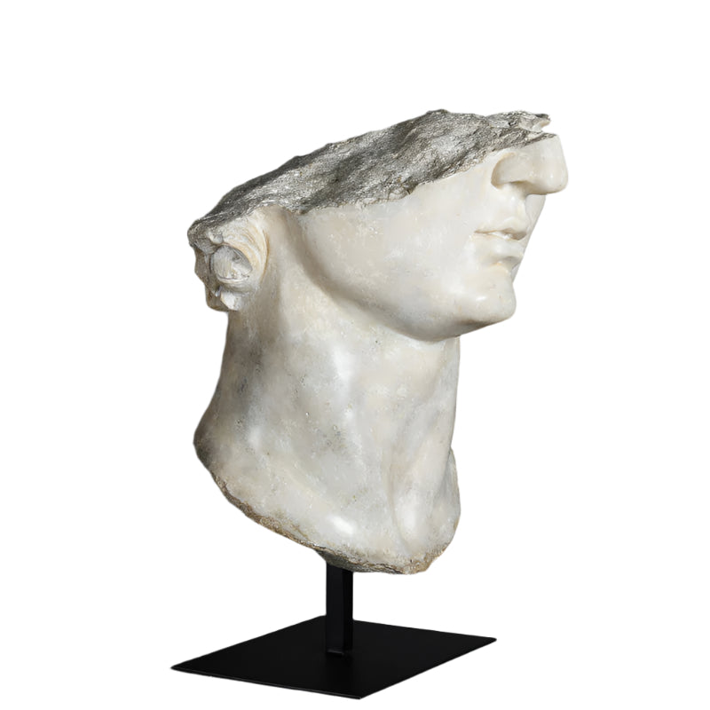 A white marble sculpture of a youthful head, likely representing Apollo, placed on a black removable stand against a grey background.