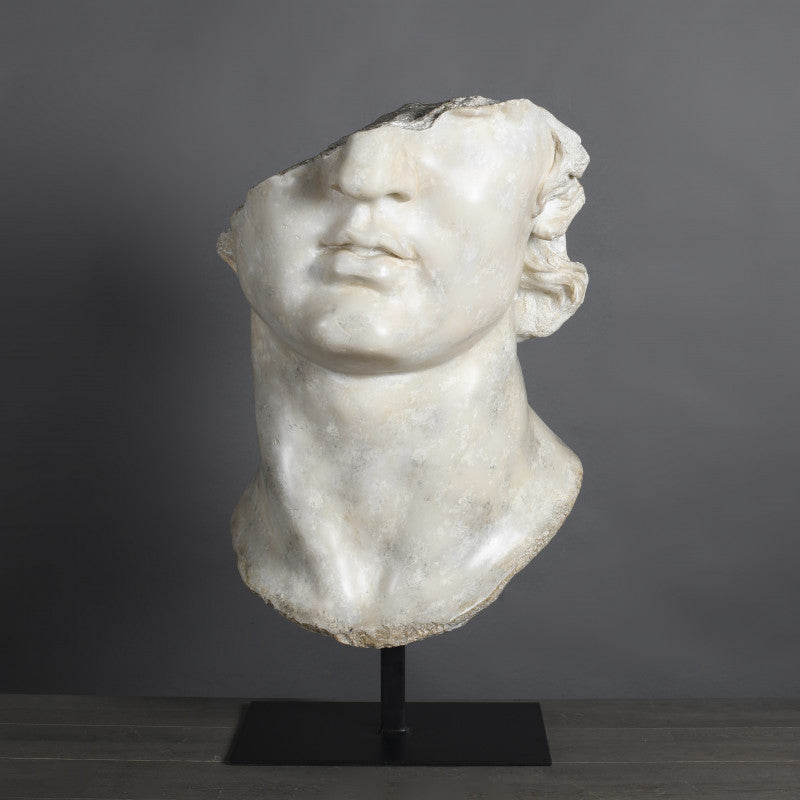 A white marble sculpture of a youthful head, likely representing Apollo, placed on a black removable stand against a grey background.